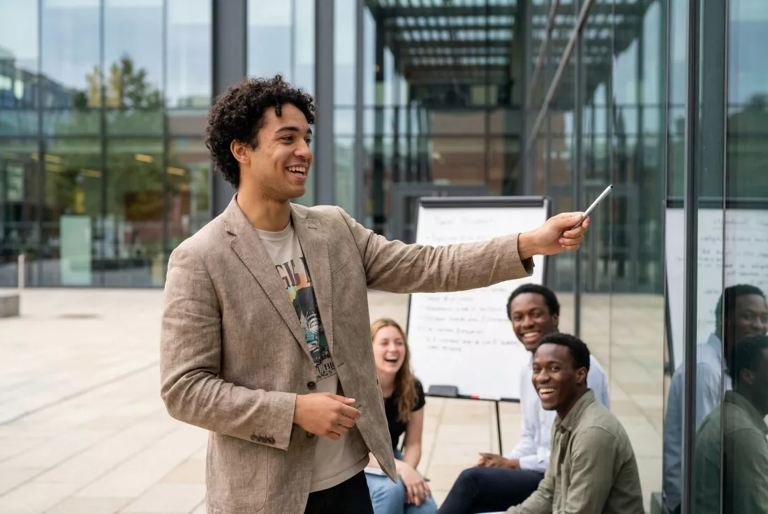A young man leads an outdoor seminar for a group of students outside a modern building.